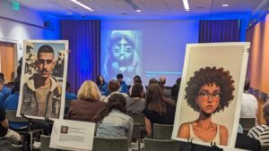 An image of a conference room with attendees facing the front of the room in rows. Toward the room three students sit with microphones in hand. The image on screen at the front is an art piece that conveys how one high school young woman felt uncomfortable with the idea of AI art, drawing an image herself. At the back of the room there are images of self-portraits students created with art.